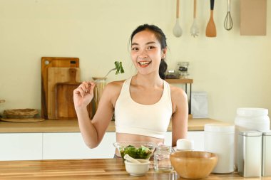Bir kadın gülümsüyor ve önünde salatayla çatal tutuyor. Masa kâseler, fincanlar ve kaşıklarla dolu. Masanın üzerinde birkaç şişe ve kavanoz var.