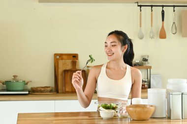 Bir kadın gülümsüyor ve önünde salatayla çatal tutuyor. Masa kâseler, fincanlar ve kaşıklarla dolu. Masanın üzerinde birkaç şişe ve kavanoz var..
