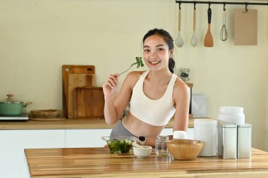 Bir kadın gülümsüyor ve önünde salatayla çatal tutuyor. Masa kâseler, fincanlar ve kaşıklarla dolu. Masanın üzerinde birkaç şişe ve kavanoz var..