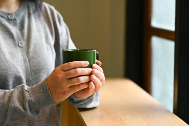 Bir kadın elinde yeşil bir kahve fincanı tutuyor. Gri bir kazak giyiyor ve bir masada oturuyor. Rahatlama ve rahatlama kavramı, kadının rahat bir ortamda kahvesinden hoşlanması gibi.