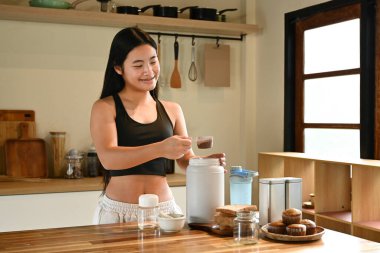 Bir kadın mutfakta protein içeceği yapıyor. Gülümsüyor ve sürecin tadını çıkarıyor..