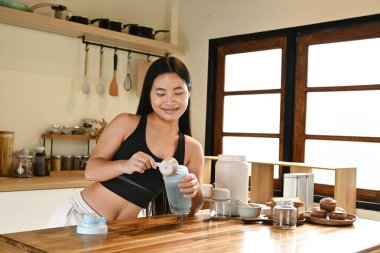 Bir kadın mutfakta protein içeceği hazırlıyor. Gülümsüyor ve sürecin tadını çıkarıyor..