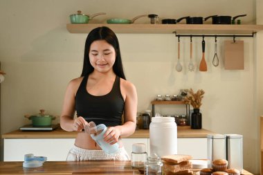 Bir kadın mutfakta protein içeceği yapıyor. Gülümsüyor ve sürecin tadını çıkarıyor.