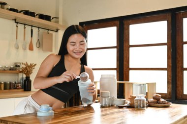 Bir kadın mutfakta protein içeceği hazırlıyor. Gülümsüyor ve sürecin tadını çıkarıyor.