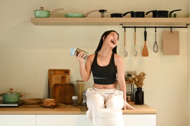 Bir kadın mutfak tezgahında elinde bir şişe protein içeceğiyle oturuyor. O gülümsüyor ve mutfakta eğleniyor. Mutfak çeşitli aletlerle donatılmıştır..