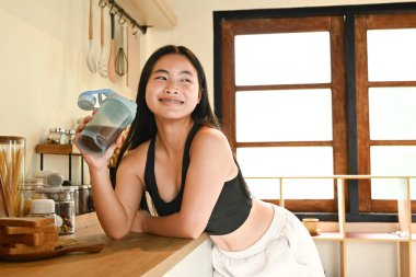 Bir kadın elinde bir bardak protein içeceğiyle mutfakta duruyor..