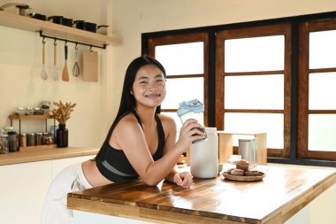 Bir kadın elinde bir bardak protein içeceğiyle mutfakta duruyor..