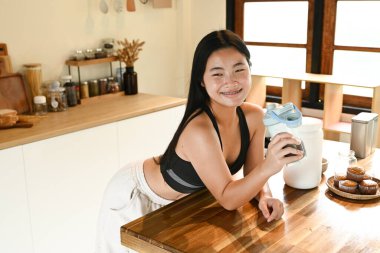 Bir kadın elinde bir bardak protein içeceğiyle mutfakta duruyor..