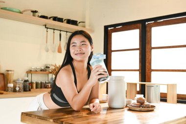 Bir kadın elinde bir bardak protein içeceğiyle mutfakta duruyor..