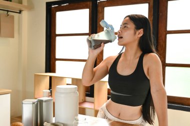 Bir kadın mutfakta smoothie içiyor. Mutfak bir fincan, bir kase, bir kaşık ve bir şişe gibi çeşitli malzemelerle dolu. Kadın bir sandalyede oturuyor..