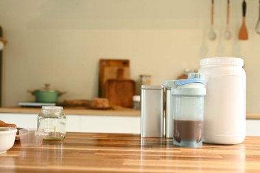 Bir kavanoz protein tozu, bir blender ve bir teneke kutunun yanındaki ahşap masada duruyor. Sahne birinin sağlıklı bir yemek ya da atıştırmalık hazırladığını gösteriyor. Mikserin varlığı....