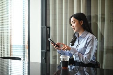 Bir kadın elinde cep telefonuyla bir masada oturuyor. Gülümsüyor ve mutlu..
