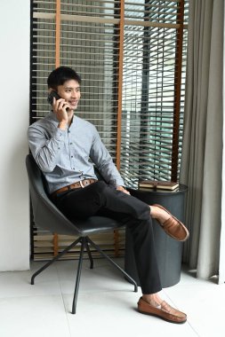 Bir adam sandalyede oturuyor ve cep telefonuyla konuşuyor. Gömlek ve pantolon giyiyor ve kahverengi bir kemeri var.