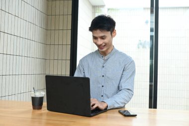 Bir adam elinde dizüstü bilgisayar ve bir fincan kahveyle masada oturuyor. Gülümsüyor ve işinden zevk alıyor.