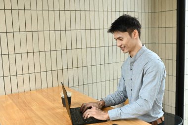 Bir adam elinde dizüstü bilgisayar ve bir fincan kahveyle masada oturuyor. Gülümsüyor ve işinden zevk alıyor..
