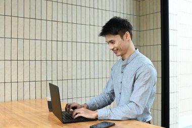 Bir adam elinde dizüstü bilgisayar ve bir fincan kahveyle masada oturuyor. Gülümsüyor ve işinden zevk alıyor..