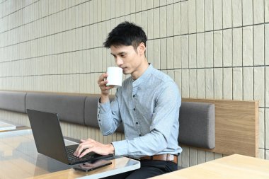Bir adam elinde dizüstü bilgisayar ve bir fincan kahveyle masada oturuyor. İşine odaklanmış ve kahvesinden zevk alıyor gibi görünüyor.