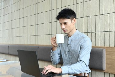 Bir adam elinde dizüstü bilgisayar ve bir fincan kahveyle masada oturuyor. İşine odaklanmış ve kahvesinden zevk alıyor gibi görünüyor..
