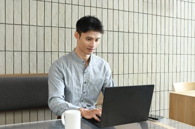 Bir adam elinde dizüstü bilgisayar ve bir fincan kahveyle masada oturuyor. Gülümsüyor ve işinden zevk alıyor..