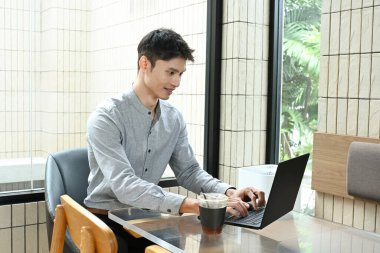 Bir adam elinde dizüstü bilgisayar ve bir fincan kahveyle masada oturuyor. O, işine odaklanmış ve ciddi bir ruh hali içinde.
