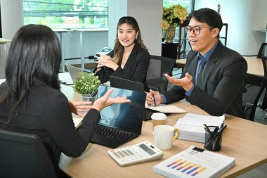 Takım elbiseli üç kişi laptopları ve kağıtları olan bir masada oturuyor. Gülümsüyorlar ve iyi vakit geçiriyor gibi görünüyorlar..