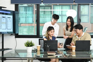 Bir grup insan dizüstü bilgisayarları ve cep telefonlarıyla bir masanın etrafında oturuyor. Hepsi cihazlarına bakıyor ve birlikte çalışıyor gibi görünüyorlar. Sahne odaklı ve işbirlikçi..