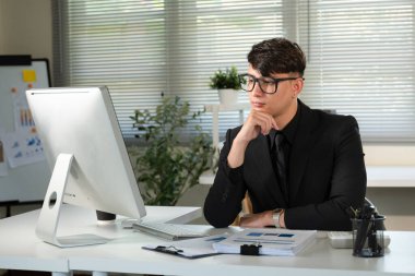 Takım elbiseli bir adam bilgisayar monitörünün önünde masada oturuyor. Gözlük takıyor ve işine odaklanmış durumda...