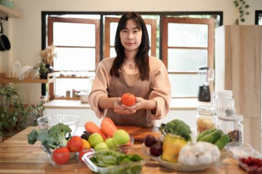 Çizgili önlüklü genç Asyalı kadın mutfakta yeşil sebze ve taze sebzelerle çevrili kırmızı bir domates tutuyor..