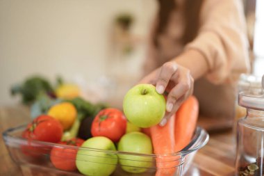 Yeşil elma tutan önlüklü Asyalı kadın. Tahta tezgahın üzerinde çeşitli sebzeler ve meyveler var. Mutfakta. Samimi..