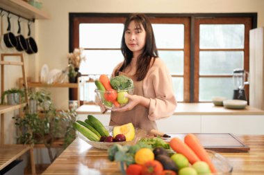 Bir kadın mutfakta bir kase sebze tutuyor. Kadın taze ürünlerle çevrili olduğu için sağlık ve refah kavramı.