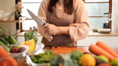 Bir kadın, havuç, domates ve brokoli de dahil olmak üzere çeşitli sebze ve meyvelerle bir masanın önünde duruyor. Elinde bıçak var ve yemek hazırlıyor..