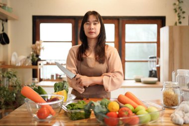 Bir kadın, havuç, domates ve brokoli de dahil olmak üzere çeşitli sebze ve meyvelerle bir masanın önünde duruyor. Elinde bıçak var ve yemek hazırlıyor.