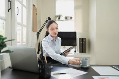 Modern ofiste elinde kahve fincanı ve dijital tablet tutan genç bir kadın. Masasında dizüstü bilgisayarı var. Rahat ve düşünceli bir ifade..