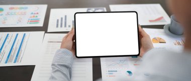 Bir kişi beyaz ekranlı bir tablet tutuyor ve birçok kağıtlı bir masaya bakıyor..