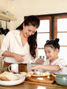 Bir anne ve bir çocuk mutfakta yemek hazırlıyorlar. Kadın gülümsüyor. Mutfak çeşitli alet ve gereçlerle dolu..