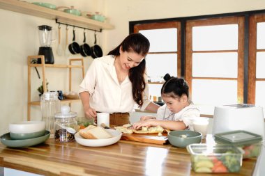 Bir anne ve bir çocuk mutfakta yemek hazırlıyorlar. Kadın gülümsüyor. Mutfak çeşitli alet ve gereçlerle dolu..