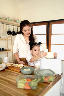 Mutlu anne kızına kahvaltı ekmeği ve ekmek kızartma makinesini evde birlikte yapmayı öğretiyor. Sevgi dolu aile. Çocuk kız heyecanlı görünüyor Anne içeride yemek yapıyor.