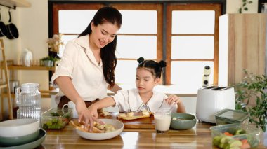 Bir anne ve bir çocuk mutfakta yemek hazırlıyorlar. Kadın gülümsüyor. Mutfak çeşitli alet ve gereçlerle dolu..