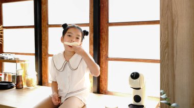 Genç bir kız pencerenin yanındaki mutfak tezgahında oturuyor ve ekmek yiyor. Mutfak çeşitli aletler ve aletlerle donatılmıştır..