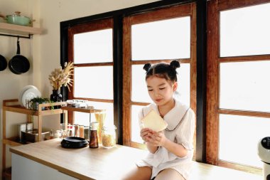 Genç bir kız pencerenin yanındaki mutfak tezgahında oturuyor ve ekmek yiyor. Mutfak çeşitli aletler ve aletlerle donatılmıştır.