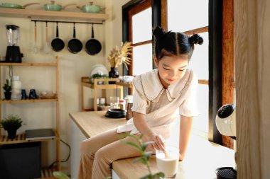 Genç bir kız pencerenin yanındaki mutfak tezgahında oturuyor ve ekmek yiyor. Mutfak çeşitli aletler ve aletlerle donatılmıştır..