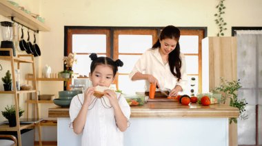 Küçük bir kız beklerken bir anne sebze kesiyor. Kız mutfakta ekmek yiyor...