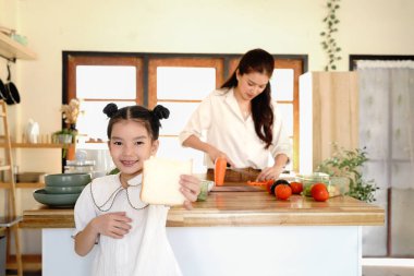 Küçük bir kız beklerken bir anne sebze kesiyor. Kız mutfakta ekmek yiyor...