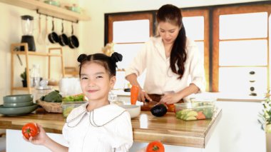 Genç bir kız annesiyle mutfakta duruyor, sebzeleri doğruyor. Kız elinde bir domates tutuyor ve annesi bir havucu kesiyor. Sahne canlı ve neşeli.