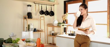 Genç kadın mutfak tezgahında duruyor, kahve içiyor ve kitap okuyor..