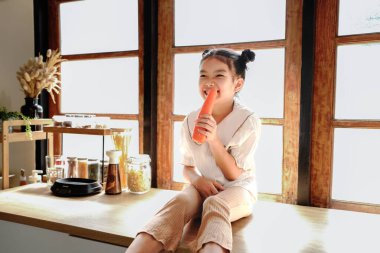 A young girl is sitting on a counter singing with a carrot. The scene is set in a kitchen with a window in the background.