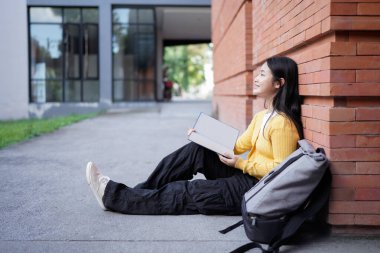 A high school girl sits on the ground her reading a book, surrounded by her backpack and books, reflecting feelings of happy on a university campus.