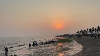 Tropikal bir plajda gün batımında insanlar toplanıyor ve altın saatin tadını çıkarıyorlar.