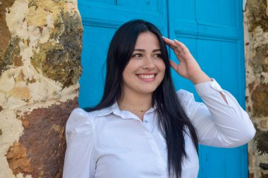 Siyah saçlı, beyaz bluzlu, gülümseyen ve elini başına koyan bir kız.