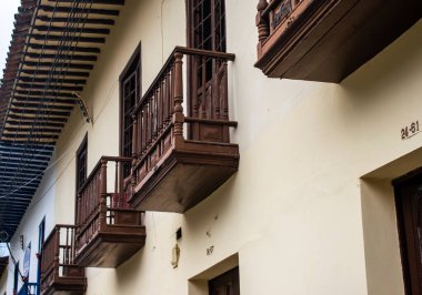 Kahverengi ahşap balkonları, fenerleri ve sarı duvarları olan eski bir koloni evi.